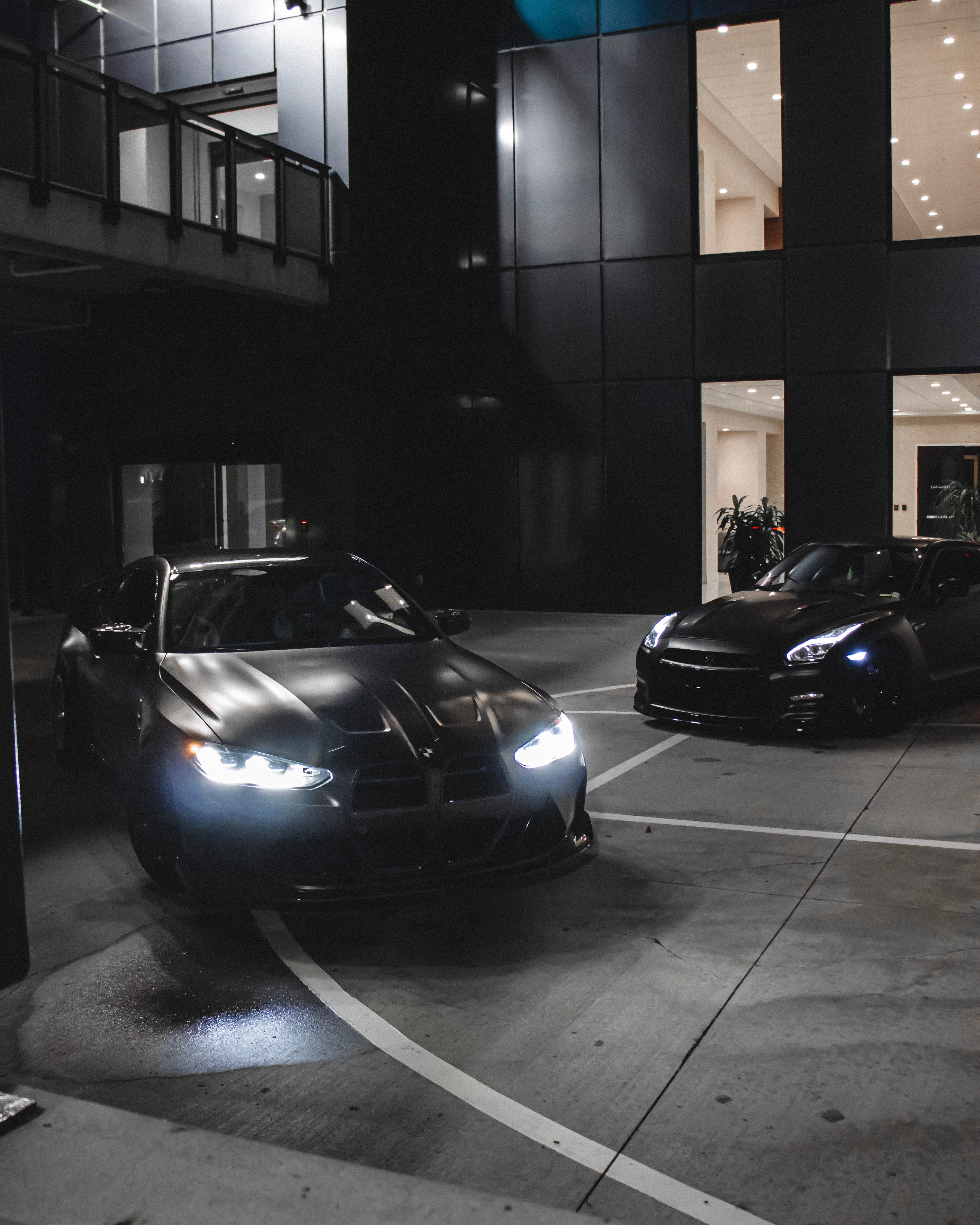 Two black sports cars parked in a parking garage with their headlights on.
